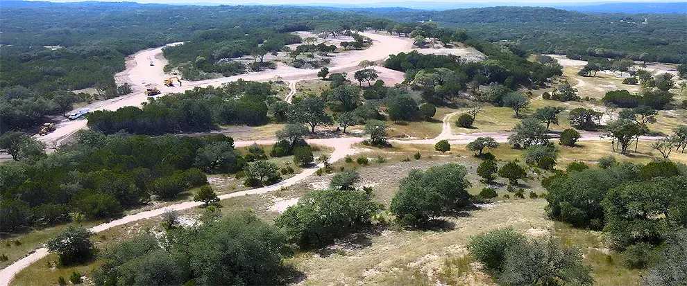 Aerial view of Texas Hill Country development under construction, showing roadway improvements, and site preparation work weaving in between the natural landscape of trees and hills.